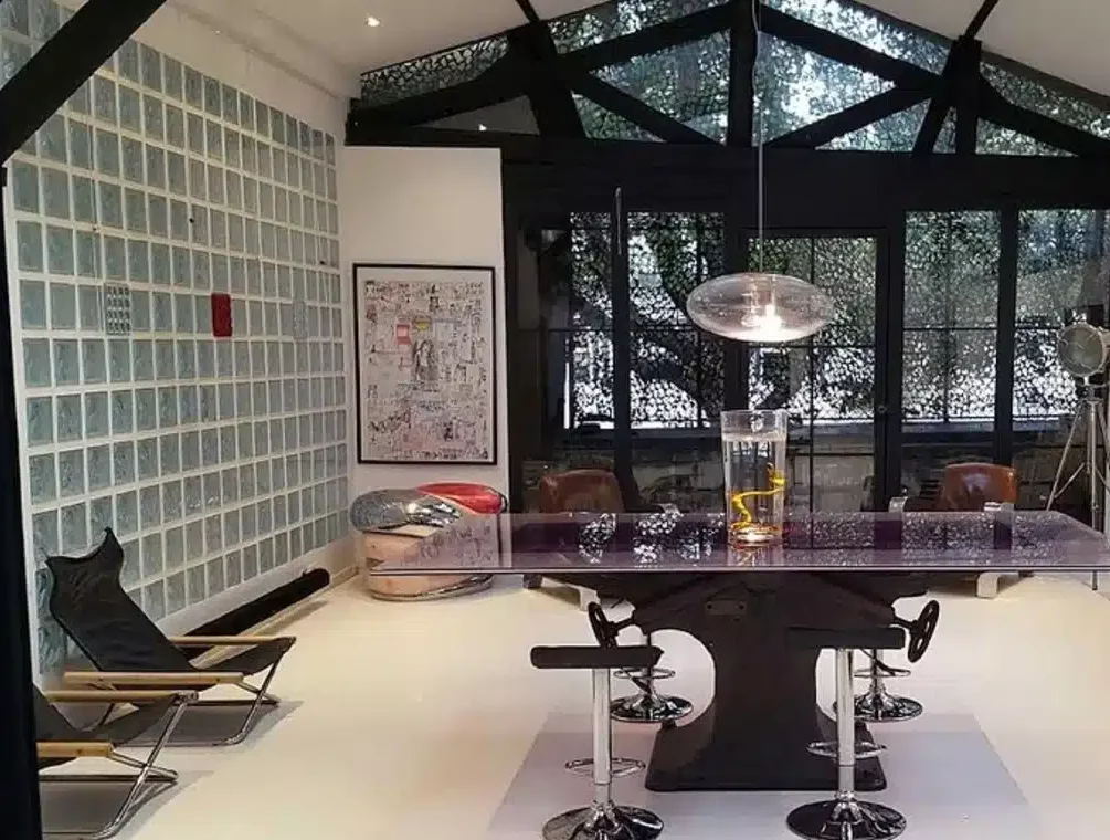 LE LOFT MONTMARTRE (75018) dispose d'une salle à manger moderne avec une table transparente, des chaises noires, un luminaire sculptural, des chaises longues, un mur de blocs de verre, des œuvres d'art encadrées et de grandes fenêtres donnant sur des arbres.