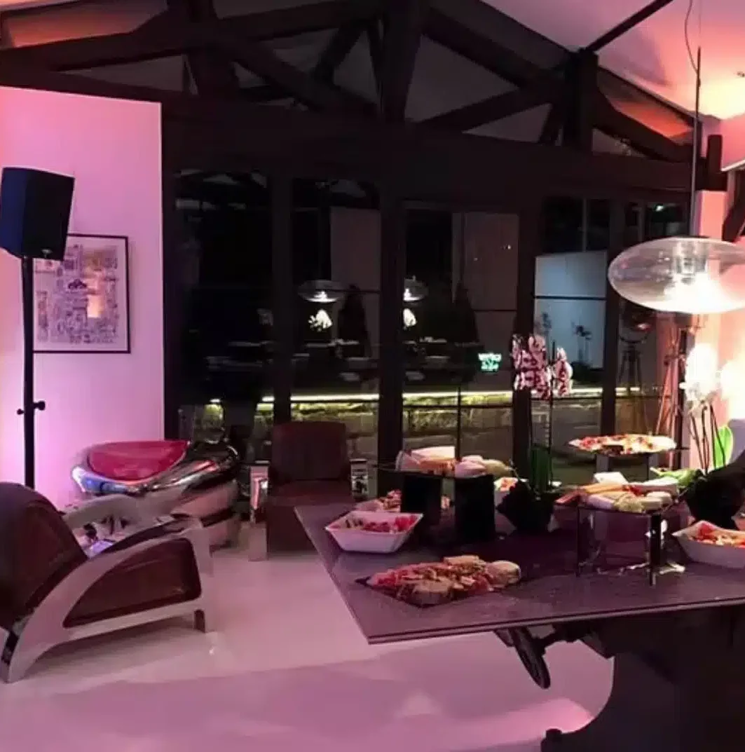 LE LOFT MONTMARTRE (75018) propose un salon moderne de nuit avec de grandes fenêtres, des éclairages roses, des chaises chics, une table dressée avec des plateaux de nourriture variés. Les lumières de la ville et les reflets sont visibles à travers les fenêtres.
