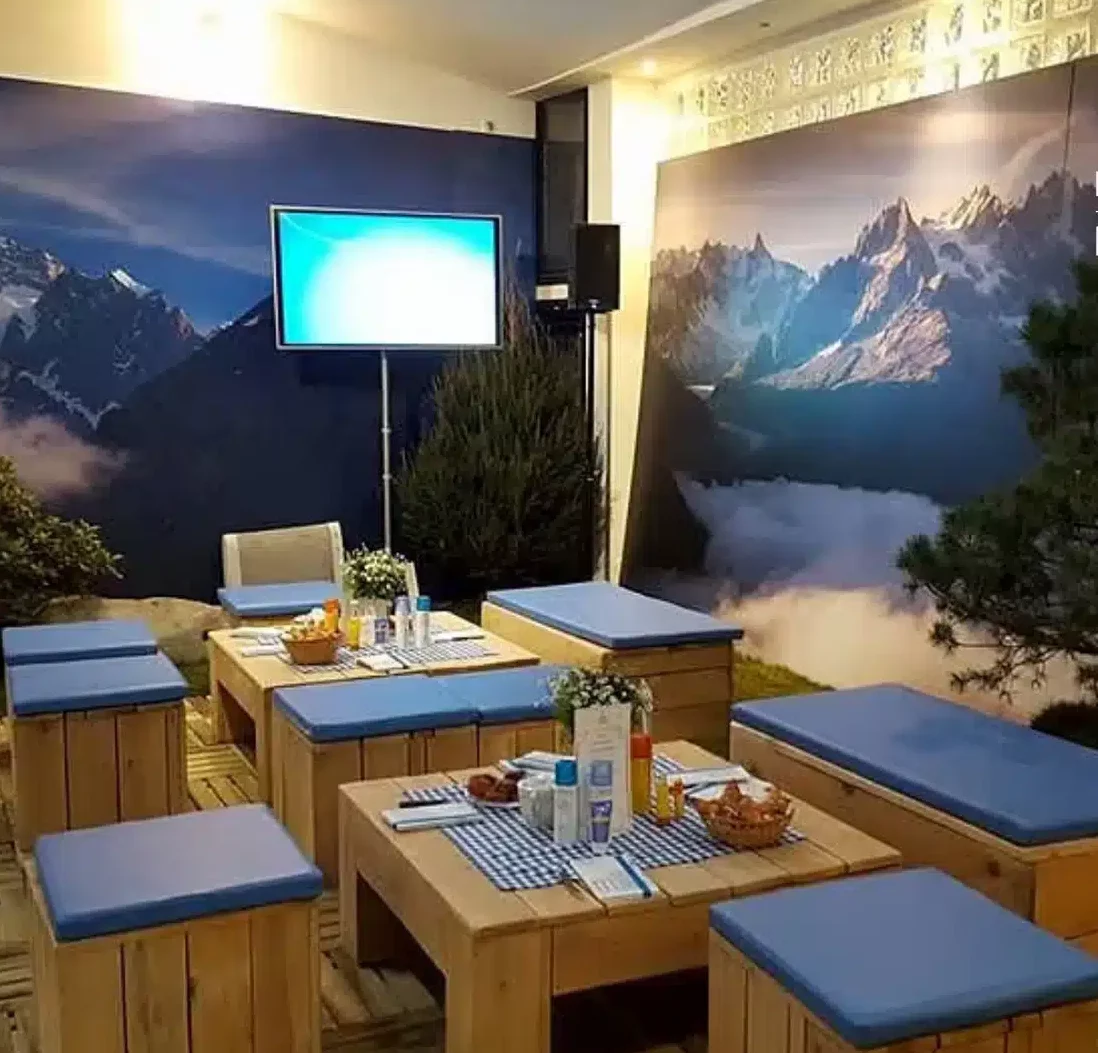 LE LOFT MONTMARTRE (75018) propose un espace intérieur confortable avec des tables en bois, des banquettes aux coussins bleus, des peintures murales de montagne sur les murs, une télévision à écran plat au-dessus d'une table, ainsi que des fleurs et des amuse-gueules sur chaque table.