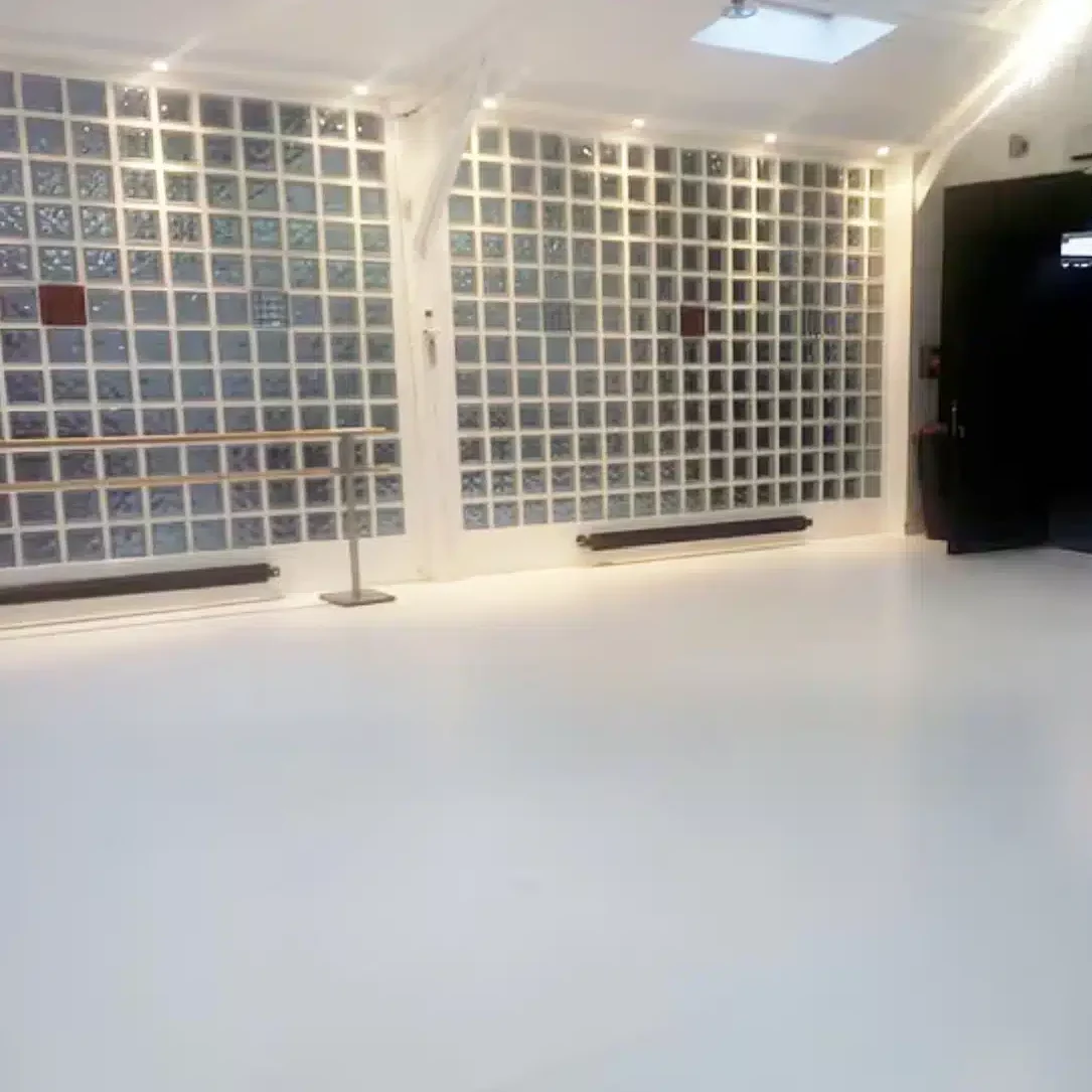 LE LOFT MONTMARTRE (75018) est un studio de danse spacieux et vide, doté d'un sol blanc, d'un mur de miroirs, d'une barre de danse et d'un éclairage zénithal. Une porte sombre apparaît sur le côté droit.