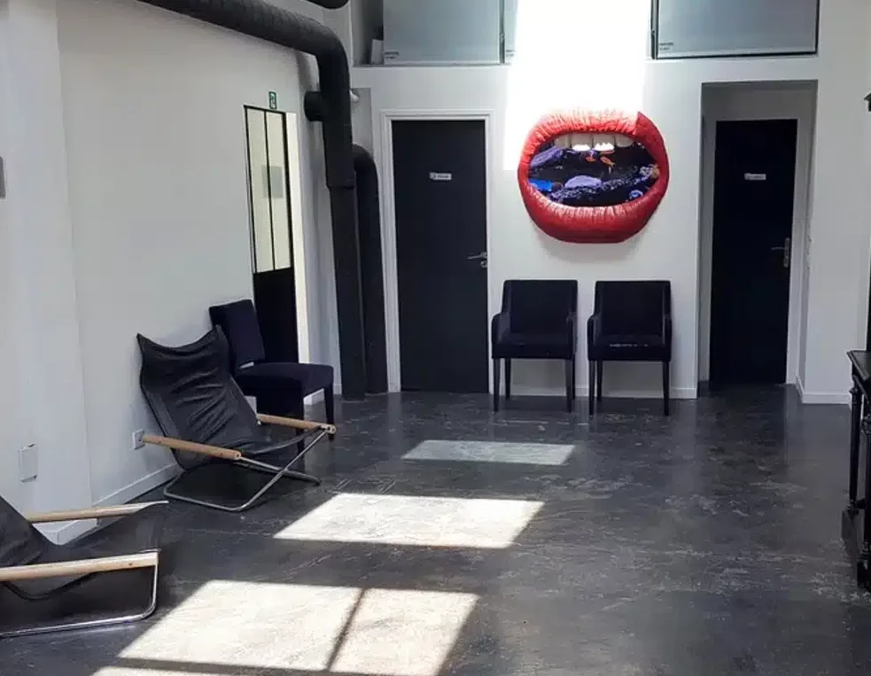 LE LOFT MONTMARTRE (75018) dispose d'une salle d'attente moderne avec des chaises noires, des sièges de salon, des portes sombres et un tableau mural représentant des lèvres rouges. La lumière du soleil passe à travers les fenêtres supérieures, illuminant les taches lumineuses sur le sol.