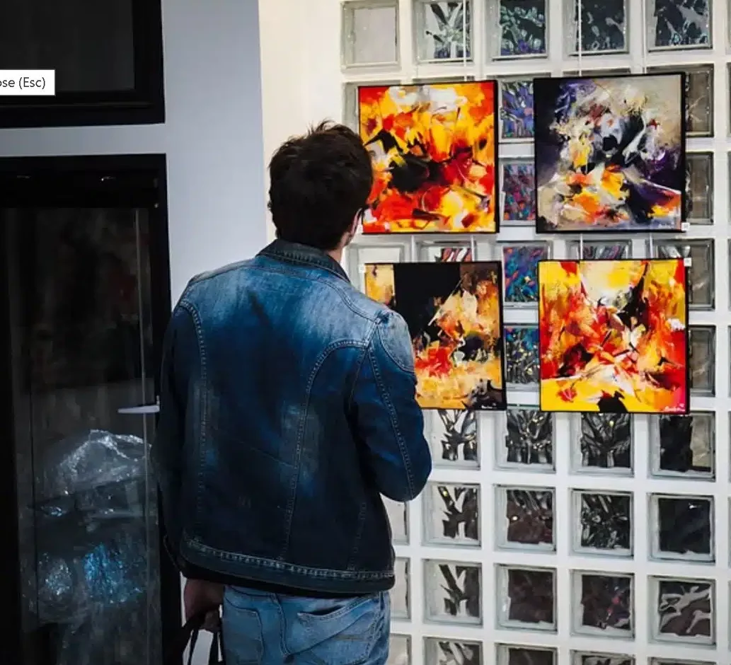 Chez LE LOFT MONTMARTRE (75018), une personne vêtue d'une veste en jean bleu admire des peintures abstraites colorées - des rouges, des jaunes et des noirs éclatants - exposées à l'intérieur contre des murs en briques de verre.