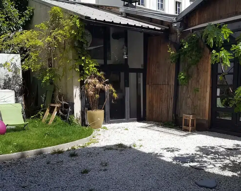 LE LOFT MONTMARTRE (75018) dispose d'une cour confortable et isolée avec du gravier et de l'herbe, entourée de bâtiments en bois et en verre, de plantes en pot, de mobilier d'extérieur et d'une végétation luxuriante.