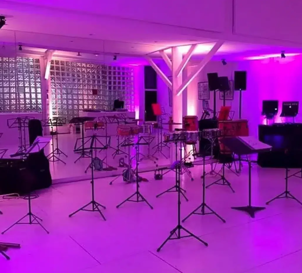 LE LOFT MONTMARTRE (75018) propose une salle de répétition de musique moderne avec un éclairage violet, des miroirs sur un mur et une disposition de pupitres, de chaises et d'instruments de percussion dans tout l'espace.