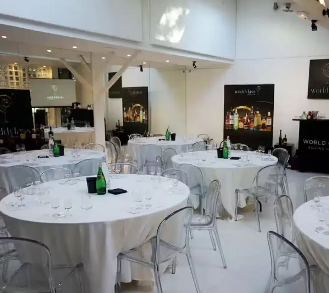 LE LOFT MONTMARTRE (75018) propose un espace événementiel lumineux avec des tables rondes drapées de blanc, des chaises claires, des verres à vin, des bouteilles vertes, des bannières de boissons noires en arrière-plan, des murs blancs et de hauts plafonds.