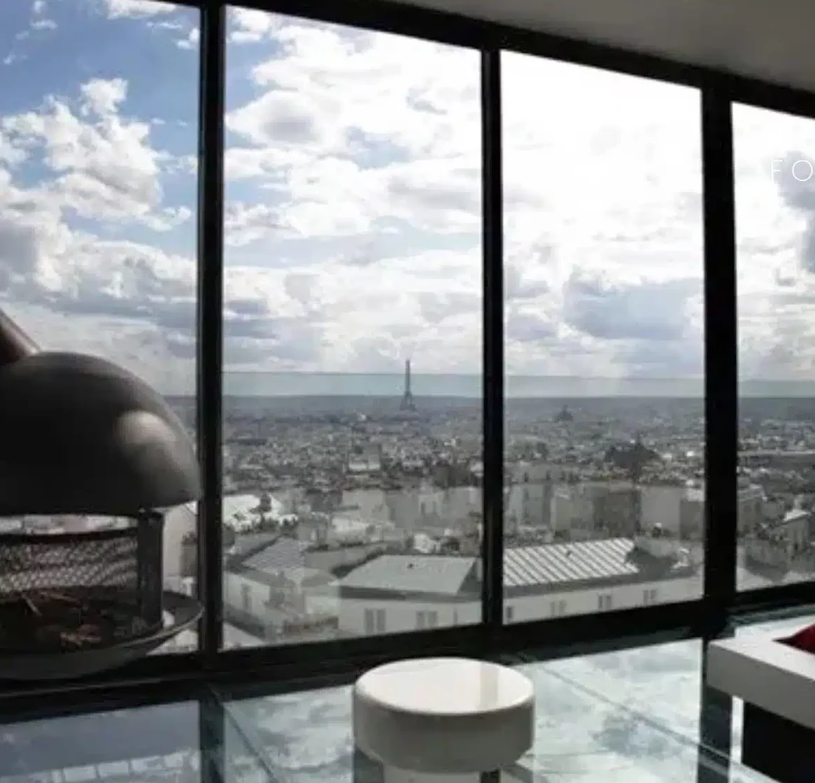 LE LOFT MONTMARTRE (75018) dispose d'un salon moderne avec des murs en verre et des vues panoramiques sur Paris, y compris la Tour Eiffel, ainsi qu'une cheminée suspendue, un sol en verre et un mobilier minimaliste.