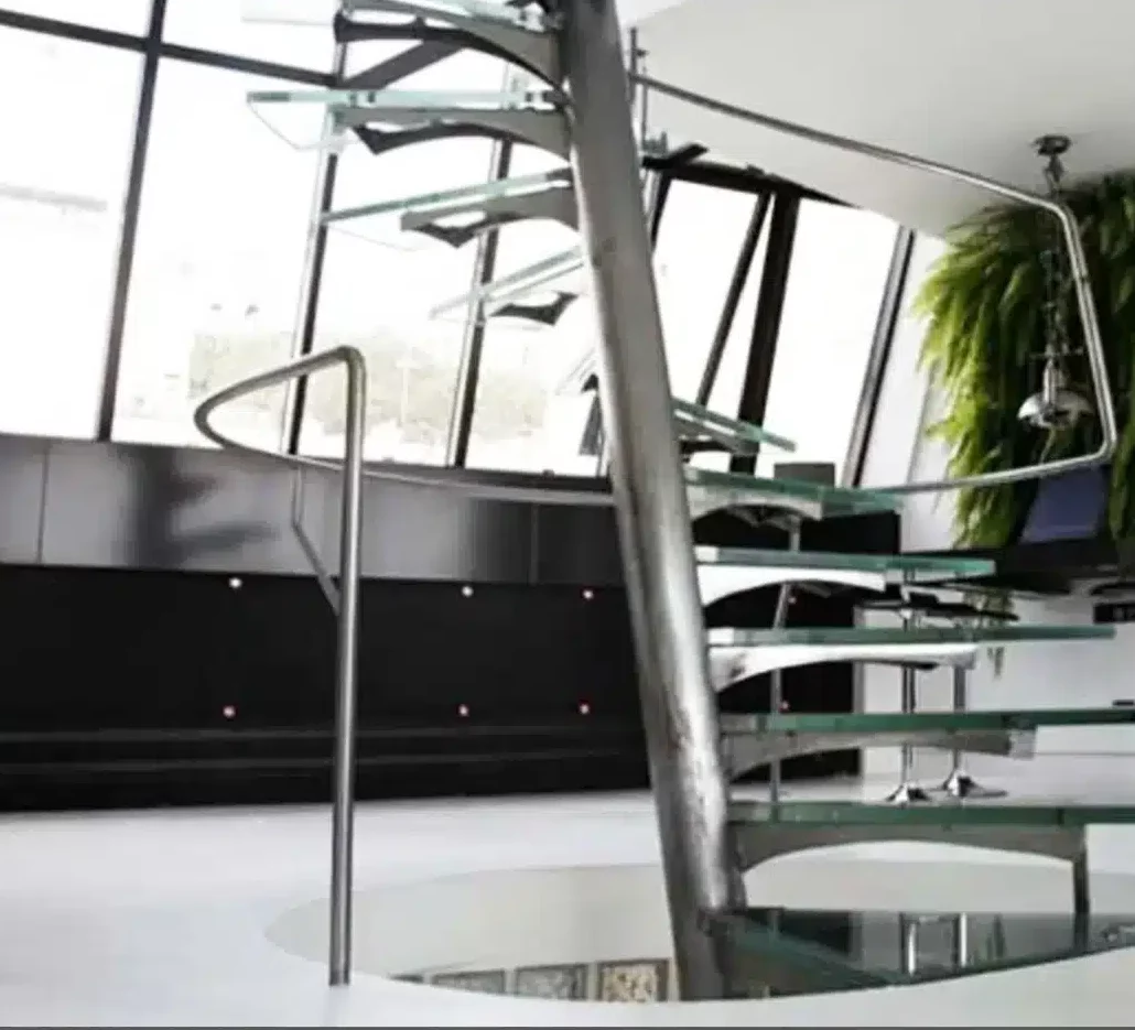Le LE LOFT MONTMARTRE (75018) présente un escalier moderne en colimaçon avec des marches en verre et une rampe en métal, éclairé par de grandes fenêtres inclinées et accentué par une plante verte suspendue près du plafond.