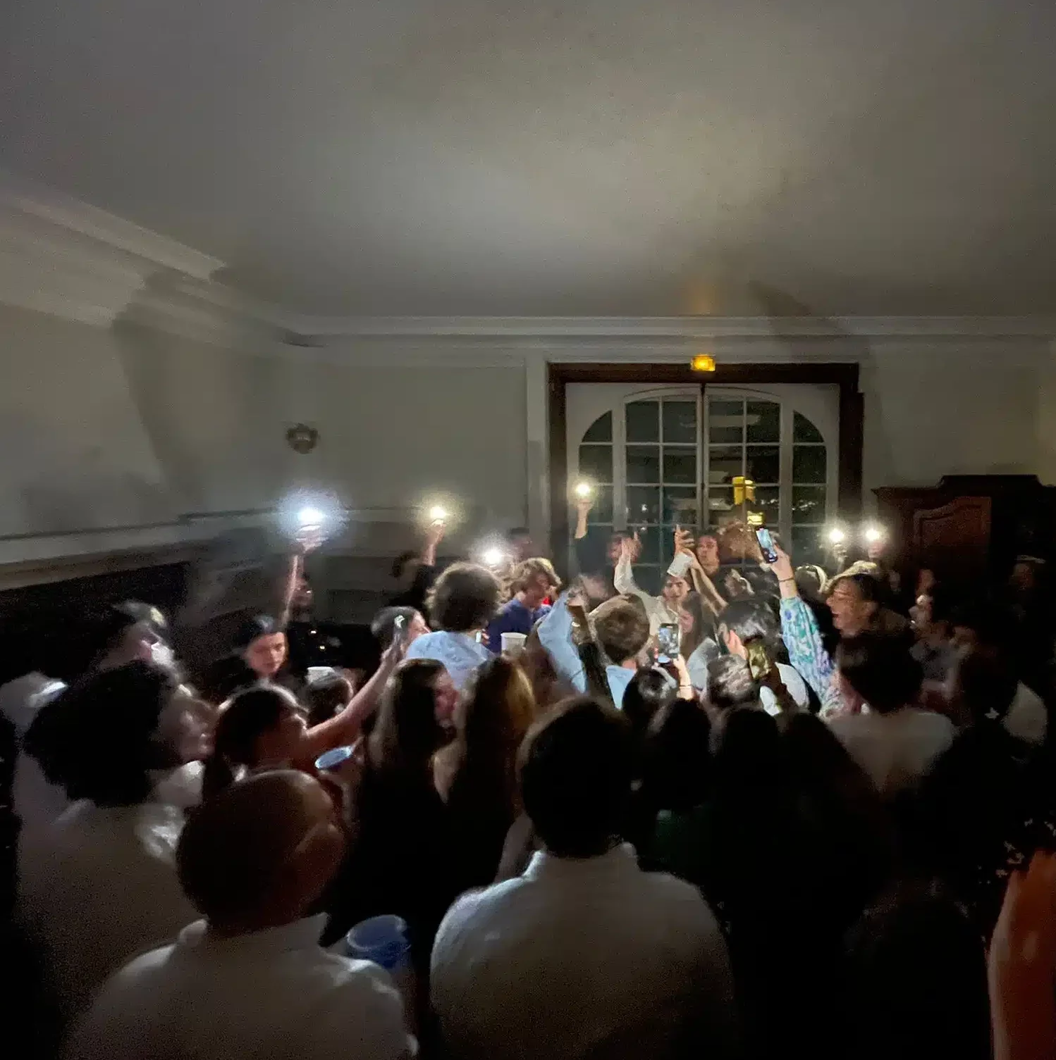 Au MANOIR DE LA CELLE SAINT CLOUD (78170), une foule participant à une fête en intérieur brandit des téléphones avec des lampes de poche, faisant la fête ensemble dans une pièce faiblement éclairée.