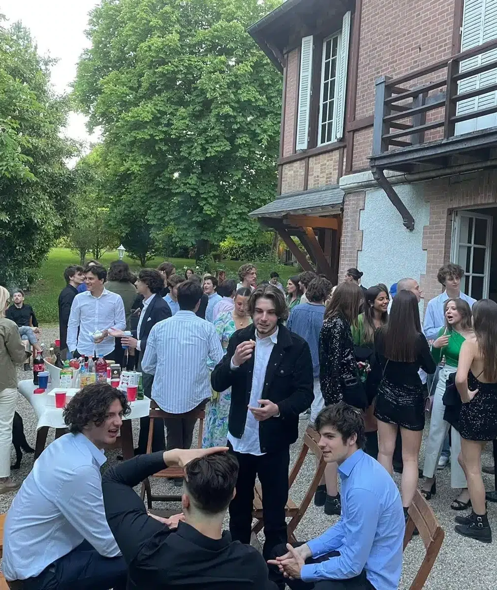 Un groupe de jeunes adultes se retrouve devant LE MANOIR DE LA CELLE SAINT CLOUD (78170). Ils discutent et se détendent autour d'une table, entourés de verdure et d'arbres luxuriants.