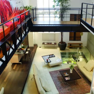 LE LOFT FACTORY (75002) est un appartement loft moderne avec une mezzanine, des balustrades en métal, des canapés blancs, une table basse en verre, des plantes en pot et de grandes fenêtres qui remplissent l'espace de lumière naturelle.