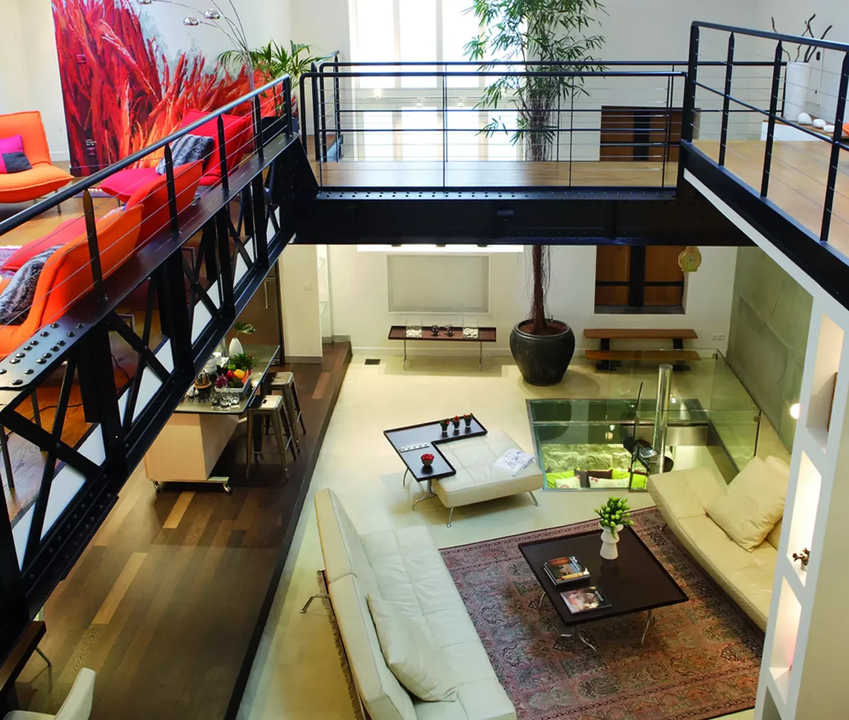 LE LOFT FACTORY (75002) est un appartement loft moderne avec une mezzanine, des balustrades en métal, des canapés blancs, une table basse en verre, des plantes en pot et de grandes fenêtres qui remplissent l'espace de lumière naturelle.