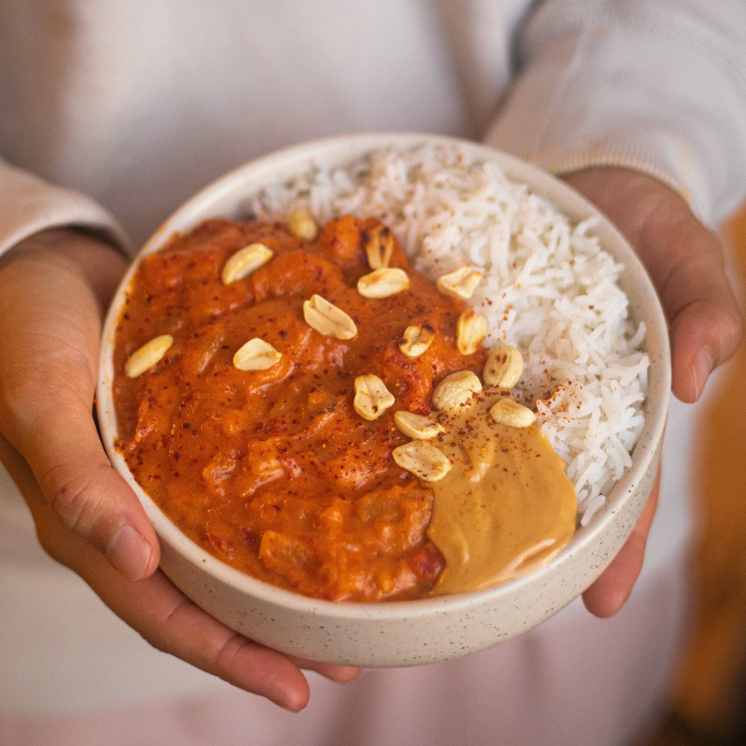 Une personne tient un bol contenant du PASTEL DE VIANDE (Barquette de 10 pièces), composé d'un ragoût crémeux à l'orange, de riz blanc, de cacahuètes et de sauce aux cacahuètes, sur un fond légèrement flou.