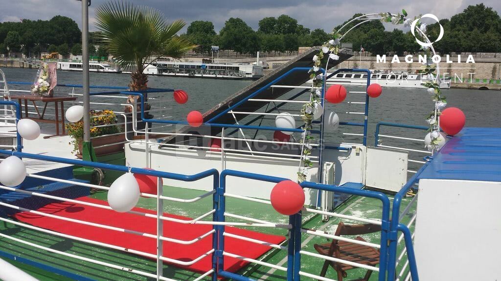 LA PÉNICHE ABYSSE (75013) présente un pont de bateau décoré de ballons rouges et blancs, d'un tapis rouge et d'une arche florale, sur une rivière avec vue sur d'autres bateaux et des arbres en arrière-plan.
