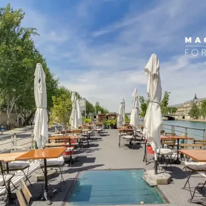 PÉNICHE LA BALLE AU BOND (75006) : Terrasse extérieure au bord de la rivière avec des tables et des chaises en bois alignées, des parapluies blancs pliés, des arbres verts bordant l'espace et un bâtiment historique en arrière-plan. Logo MAGNOLIA FOR EVENT en haut à droite.