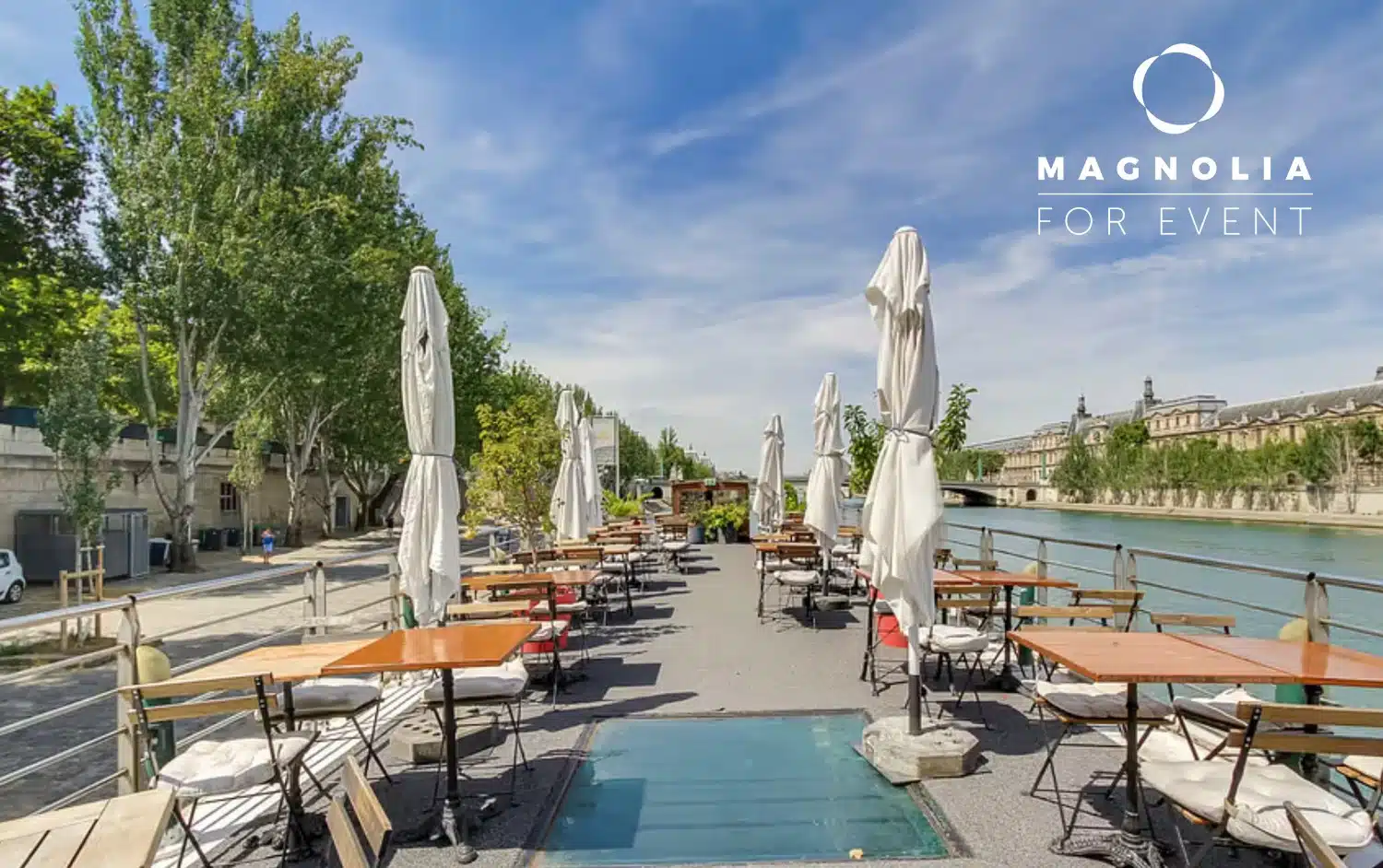 PÉNICHE LA BALLE AU BOND (75006) : Terrasse extérieure au bord de la rivière avec des tables et des chaises en bois alignées, des parapluies blancs pliés, des arbres verts bordant l'espace et un bâtiment historique en arrière-plan. Logo MAGNOLIA FOR EVENT en haut à droite.