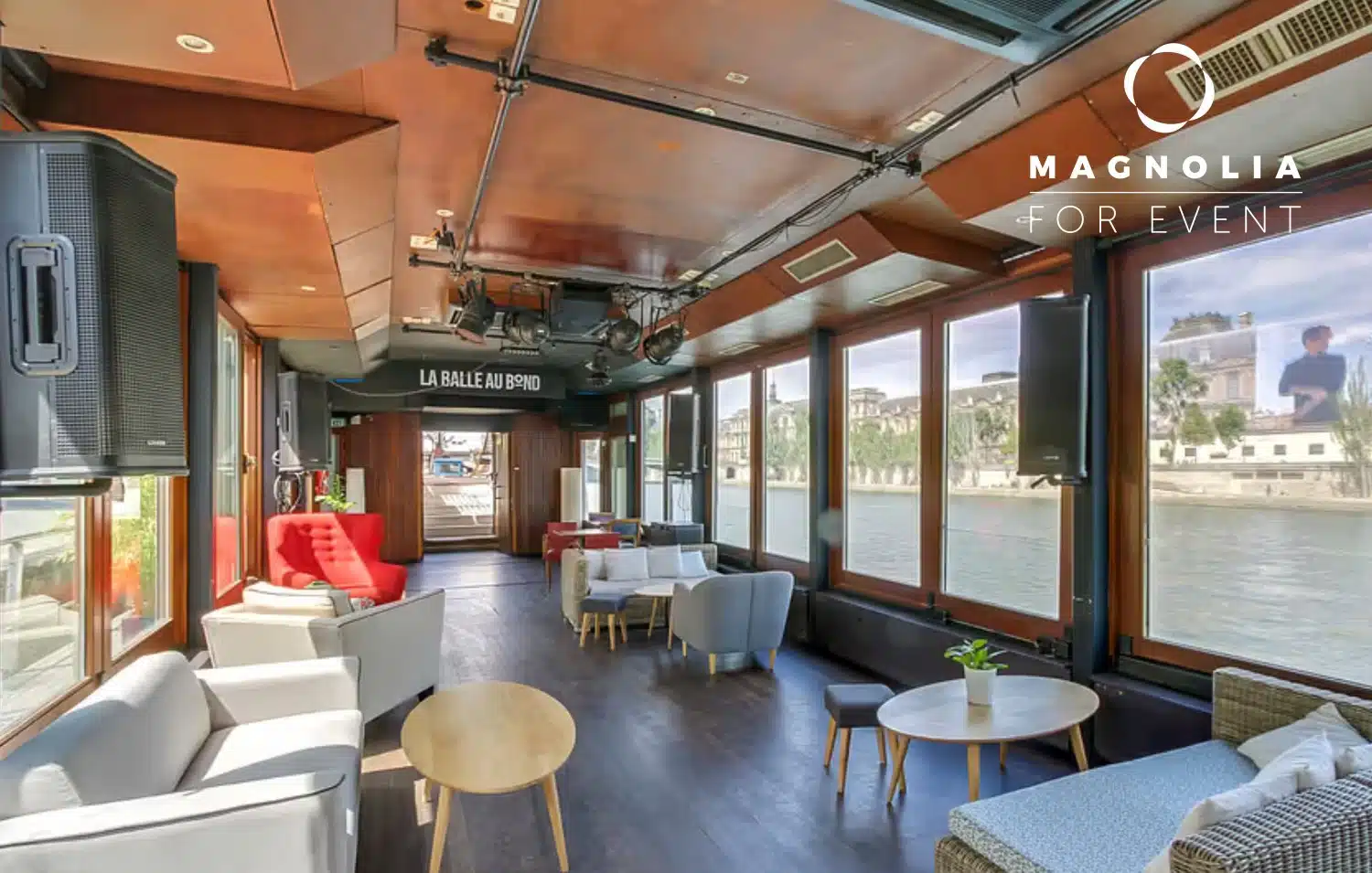 L'espace événementiel moderne PÉNICHE LA BALLE AU BOND (75006) dispose de grandes fenêtres avec vue sur la rivière, de chaises confortables, de canapés et de tables remplies de lumière naturelle. Le logo Magnolia For Event apparaît dans le coin supérieur droit.