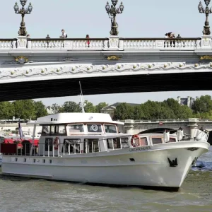 LA PÉNICHE CACHEMIRE (75004), un bateau blanc, navigue sur une rivière sous un pont orné de lampadaires, tandis que des gens marchent au-dessus. Des arbres verts et des bâtiments sont visibles à l'arrière-plan sous un ciel clair.