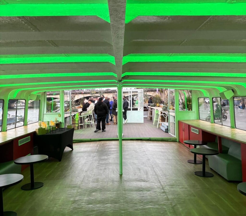 L'image montre l'intérieur de LA PÉNICHE CROISIÈRES DE BERCY (75013) avec des poutres de plafond éclairées en vert, des tables et des bancs vides, et des personnes debout à l'extérieur sur le pont au bord d'une rivière ou d'un canal.