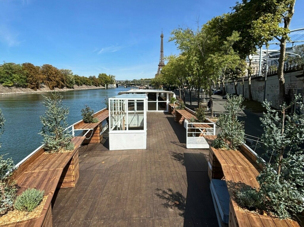 LA PÉNICHE CROISIÈRES DE BERCY (75013) représente un pont en bois avec des bancs et des plantes sur un bateau amarré au bord de la Seine à Paris, avec la Tour Eiffel visible en arrière-plan sous un ciel bleu clair.