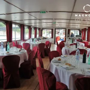 Salle à manger élégante sur le bateau LA GUEPE BUISSONNIERE (75019), avec de grandes fenêtres, des tables rondes avec des nappes blanches, des chaises rouges, de la verrerie et des bouteilles. Le logo Magnolia for Event est visible dans le coin supérieur droit.