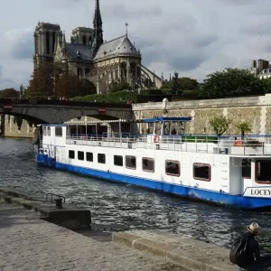 Un bateau fluvial nommé LA PÉNICHE LOCET (75013) navigue sur la Seine à Paris, avec la cathédrale Notre-Dame en arrière-plan et une personne assise sur les marches au premier plan.