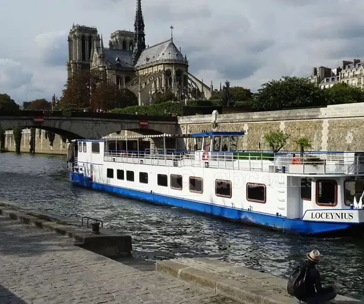Un bateau fluvial nommé LA PÉNICHE LOCET (75013) navigue sur la Seine à Paris, avec la cathédrale Notre-Dame en arrière-plan et une personne assise sur les marches au premier plan.
