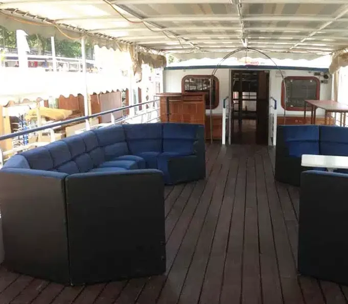 LA PÉNICHE LOCET (75013) dispose d'un pont couvert avec des planchers en bois foncé, des sièges sectionnels à coussins bleus, des tables et un espace arrière partiellement fermé. Le bateau est amarré près d'arbres et d'une clôture en bois.