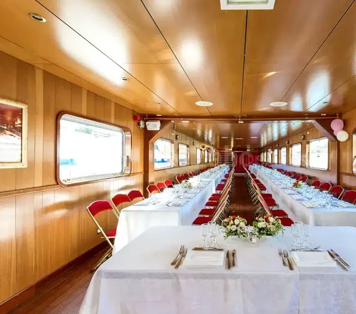 LA PÉNICHE LOCET (75013) propose une salle à manger spacieuse, lambrissée sur un bateau, avec de grandes fenêtres, deux rangées de tables avec des nappes blanches, des chaises rouges et des centres de table fleuris, parfaits pour les événements formels.