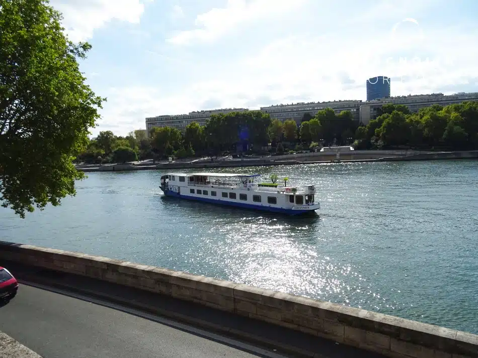 Le bateau LA PÉNICHE LOCET (75013), blanc et bleu, navigue sur une rivière calme bordée d'arbres et d'une digue en pierre, avec des bâtiments urbains et de la verdure en arrière-plan sous un ciel lumineux.