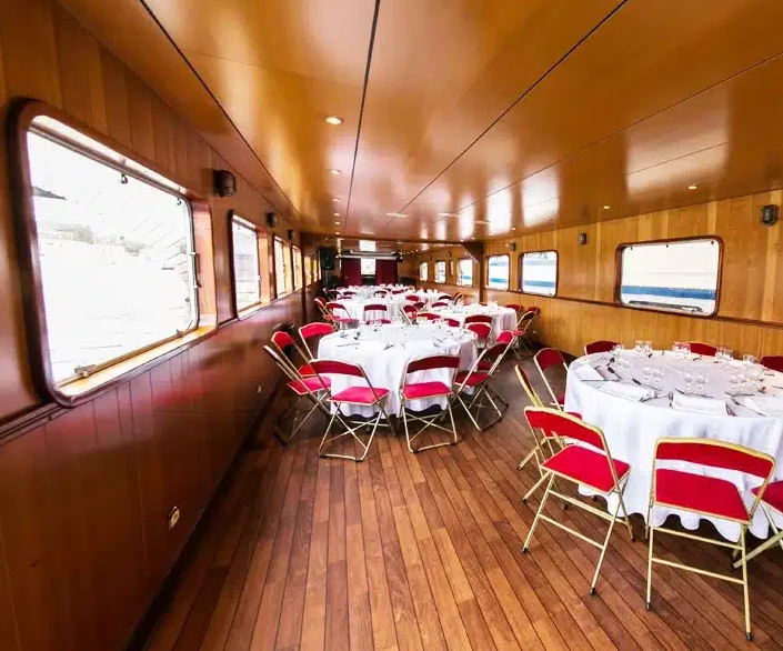 LA PÉNICHE LOCET (75013) dispose d'une grande salle boisée avec des tables rondes drapées de blanc, des chaises rouges et de grandes fenêtres rectangulaires, un lieu idéal pour les événements en bateau.
