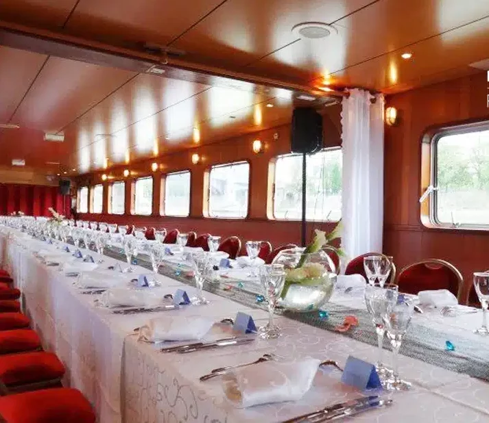 Un banquet formel est organisé sur le LA PÉNICHE LOCET (75013) avec des nappes blanches, des chaises rouges, des verres et des serviettes, entouré de grandes fenêtres latérales qui remplissent le navire de lumière naturelle.