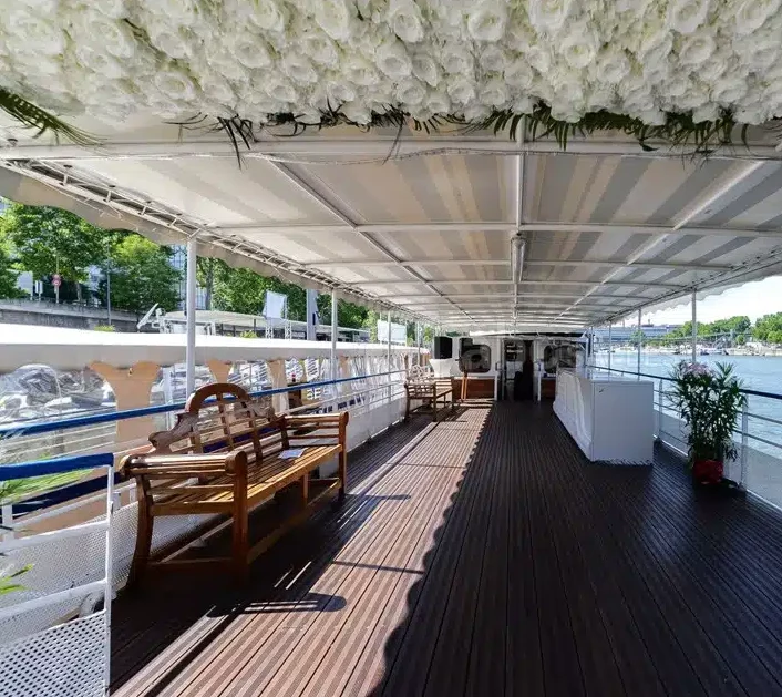 LA PÉNICHE LOCET (75013) dispose d'un pont de bateau spacieux avec un plancher en bois, un décor floral blanc au-dessus, des bancs et des places assises le long des côtés, et une vue imprenable sur la rivière et les arbres verts sous un ciel ensoleillé.