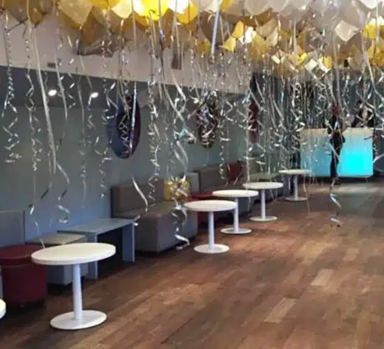 La PÉNICHE NOTRE DAME DE PARIS (75005) est dotée d'un plancher en bois, de tables rondes blanches, de sièges gris et de ballons dorés et argentés avec des rubans bouclés suspendus au plafond pour une ambiance festive.
