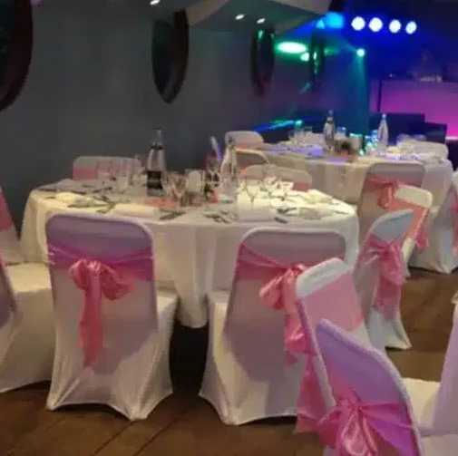 LA PÉNICHE NOTRE DAME DE PARIS (75005) propose des tables rondes avec des nappes blanches et des chaises drapées de tissus blancs noués par des écharpes roses, le tout sur fond de lumières colorées pour une ambiance festive.