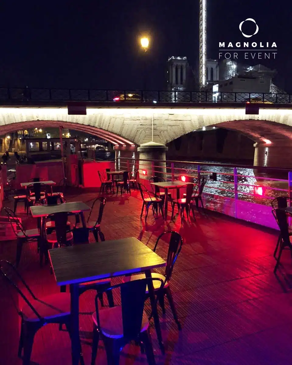 Vue nocturne de tables et de chaises vides sur le pont de LA PÉNICHE NOTRE DAME DE PARIS (75005) au bord d'une rivière, éclairée par des lumières rouges et bleues, face à un pont de pierre doucement illuminé. Le logo Magnolia For Event apparaît dans le coin supérieur droit.