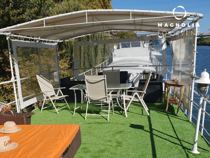 L'espace extérieur de LA PÉNICHE SÉRÉNITÉ (92390) est composé d'une table, de chaises, d'un canapé et d'un transat sous un auvent. De l'eau et des arbres se trouvent à proximité ; le logo Magnolia for Event apparaît dans le coin supérieur droit.