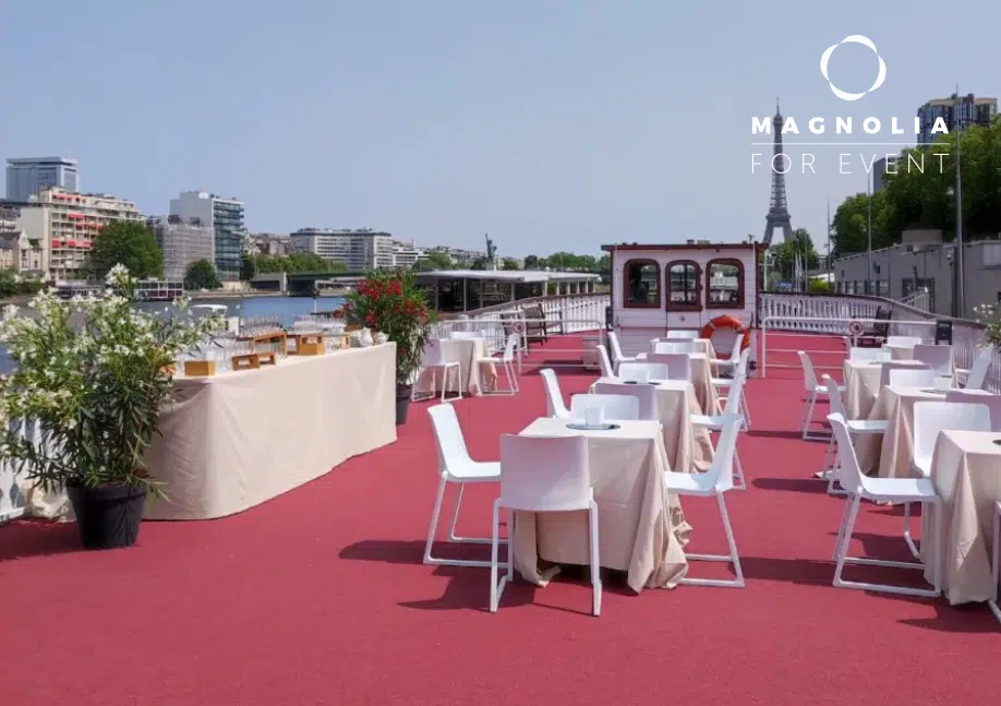 Configuration d'un événement en plein air au LE NASHVILLE TENNESSE (75015) : terrasse au bord de l'eau recouverte d'un tapis rouge avec des tables nappées de blanc, des chaises blanches, des tables de buffet, des plantes et une vue sur le paysage urbain. Logo Magnolia For Event visible dans le coin.