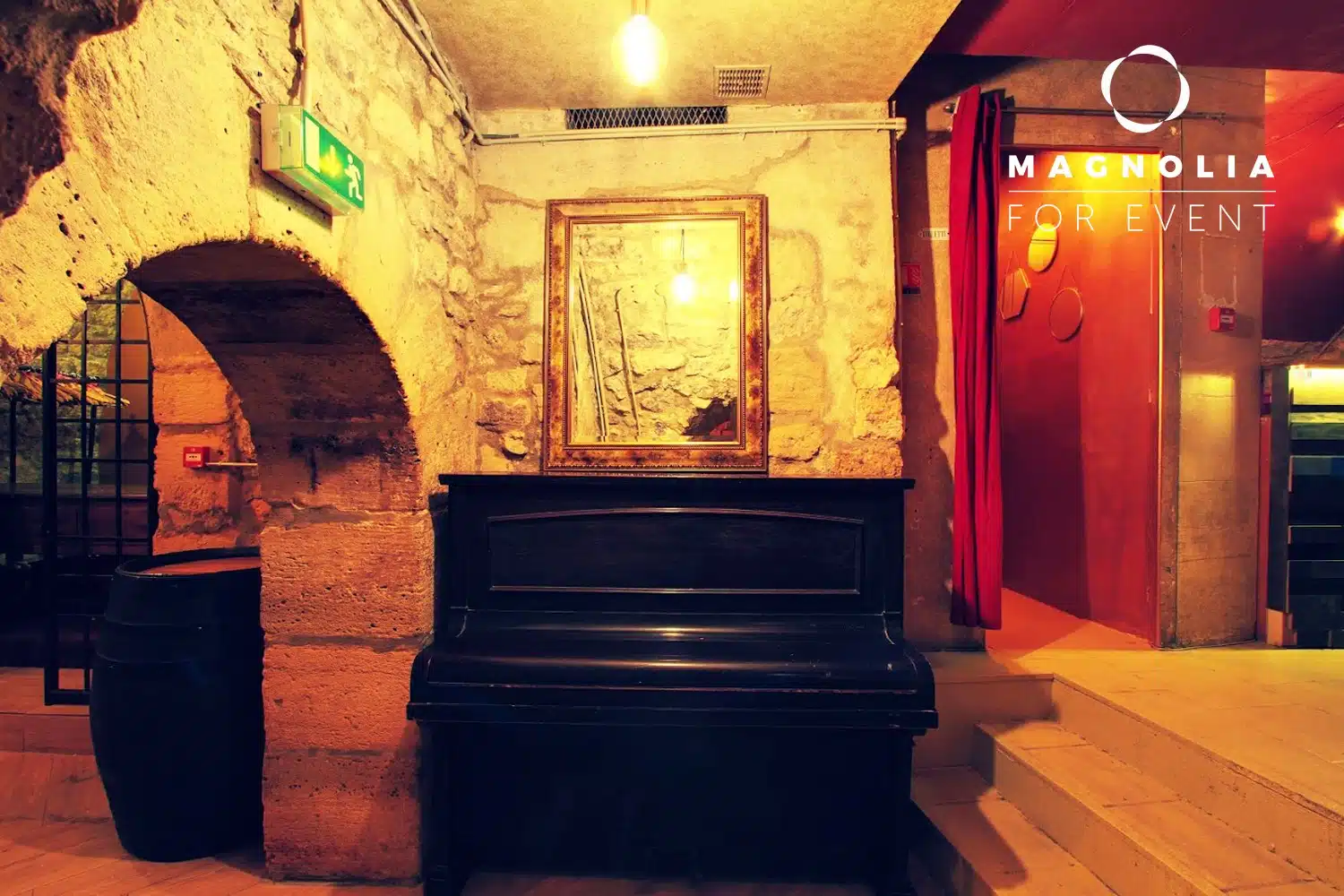 LE PETIT PARIS CACHÉ (75004) offre un espace confortable et rustique avec des murs en pierre, un piano droit noir sous un miroir, des marches à proximité et un rideau rouge, ainsi qu'un panneau de sortie lumineux. L'image de marque de Magnolia for Event apparaît dans le coin supérieur droit.