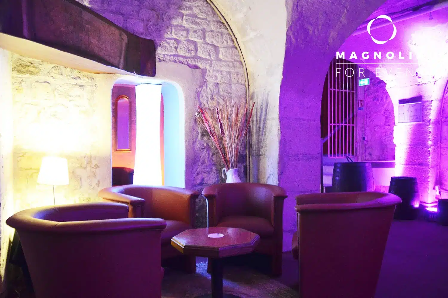 Le salon confortable du PETIT PARIS CACHÉ (75004) dispose de chaises en cuir rouge et d'une petite table dans une pièce aux murs de pierre, éclairée par une lumière douce violette et jaune, avec "Magnolia For Events" projeté sur le mur.