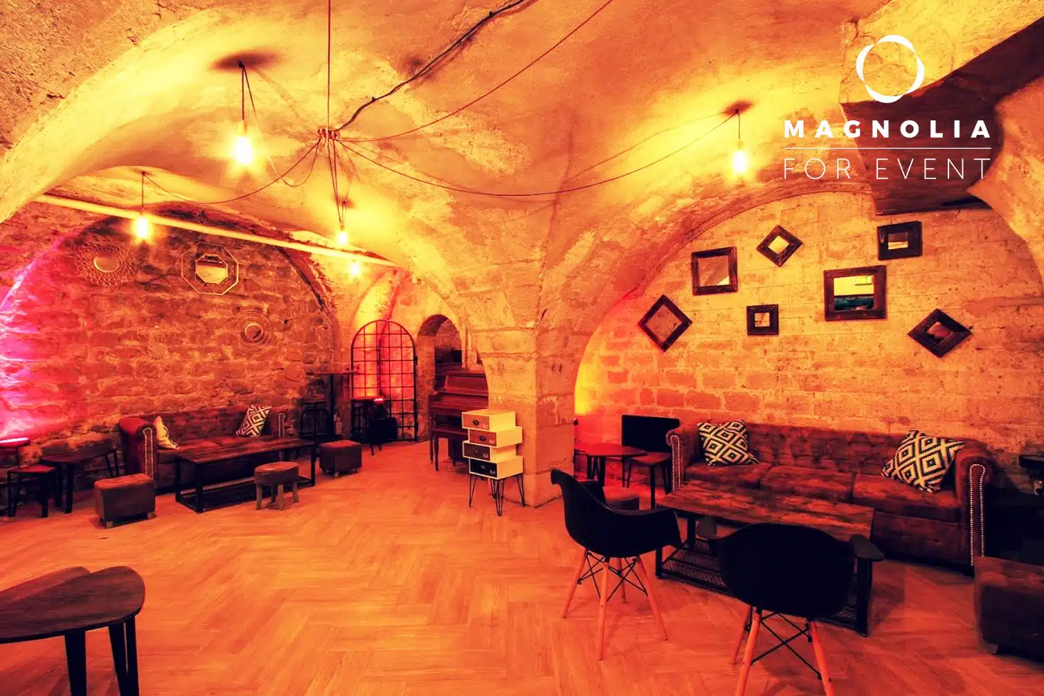 Une salle aux murs de pierre chaudement éclairée avec des plafonds voûtés, un mobilier moderne et un décor encadré, aménagée pour un événement confortable au PETIT PARIS CACHÉ (75004), avec le logo Magnolia For Event dans le coin.