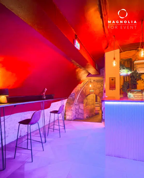LE PETIT PARIS CACHÉ (75004) se caractérise par un éclairage rose et orange vibrant, des tabourets hauts sur un comptoir épuré, des murs en briques apparentes, des luminaires suspendus et son logo dans le coin supérieur droit pour une ambiance de bar élégante.
