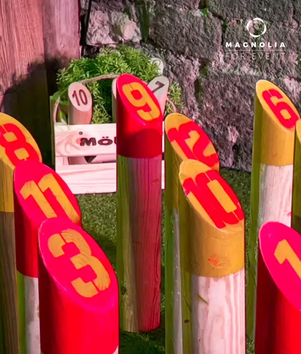 Le set LE PETIT PARIS CACHÉ (75004) présente des épingles en bois avec des chiffres rouges et jaunes sur de l'herbe, avec une boîte en bois contenant une plante à proximité. La scène extérieure est adossée à un mur de pierre.