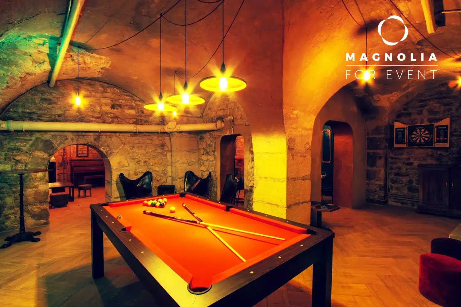 LE PETIT PARIS CACHÉ (75004) : Une salle de jeu confortable avec des murs en pierre et des plafonds voûtés, une table de billard rouge, des queues de billard, des lumières chaudes suspendues et une cible de fléchettes sur le mur du fond. Le logo de Magnolia For Event apparaît en haut à droite.