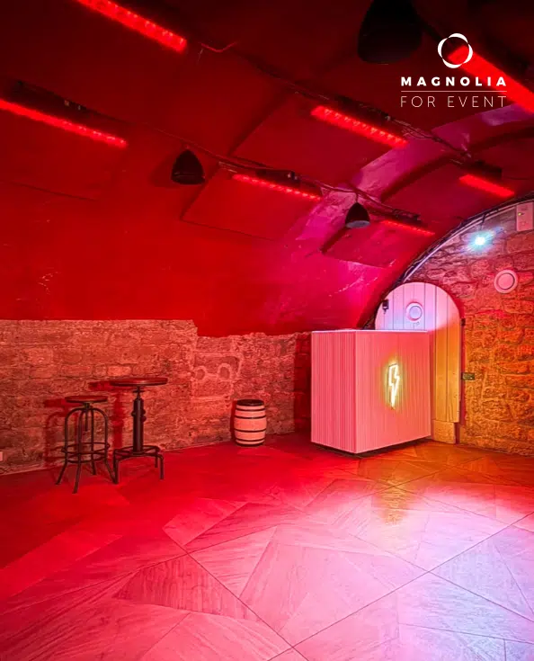 LE PETIT PARIS CACHÉ (75004) est un lieu animé avec des murs en pierre, un plafond voûté, un éclairage rouge, des tables hautes et des tabourets, un tonneau et un bar blanc avec un éclair lumineux. Le logo de Magnolia For Event apparaît dans le coin.