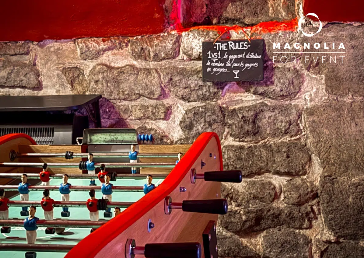 Un baby-foot aux bords rouges est posé devant un mur de pierre. Au-dessus, un petit tableau affiche les règles du jeu, et le logo "LE PETIT PARIS CACHÉ (75004)" apparaît sur le mur dans le coin supérieur droit.