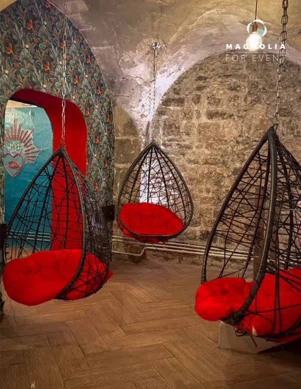 Trois fauteuils LE PETIT PARIS CACHÉ (75004) de couleur noire avec des coussins rouge vif sont suspendus à un plafond en pierre sur un sol en bois, avec une peinture murale de cœurs colorés sur le mur à motifs à l'arrière-plan.