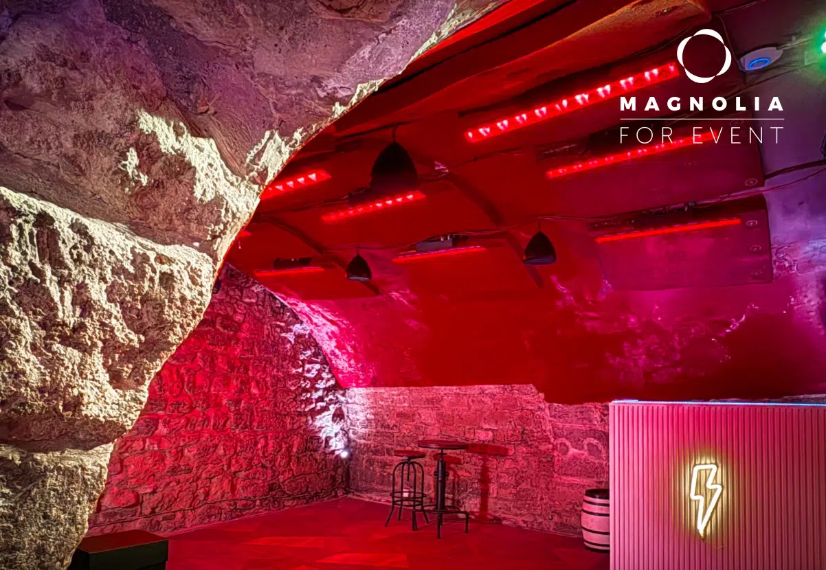 LE PETIT PARIS CACHÉ (75004) est un espace événementiel aux murs de pierre, semblable à une grotte, avec un éclairage rouge, une petite table pour deux personnes, un tonneau et une enseigne en forme d'éclair au néon. Le logo Magnolia For Event apparaît dans le coin supérieur droit.