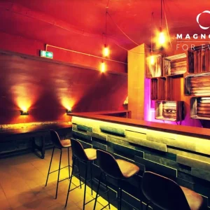 LE PETIT PARIS CACHÉ (75004) dispose d'un bar moderne avec un comptoir en bois, trois tabourets de bar noirs, un éclairage chaleureux, des œuvres d'art murales abstraites et une ambiance chaleureuse et accueillante. Le logo Magnolia For Event se trouve dans le coin supérieur droit.