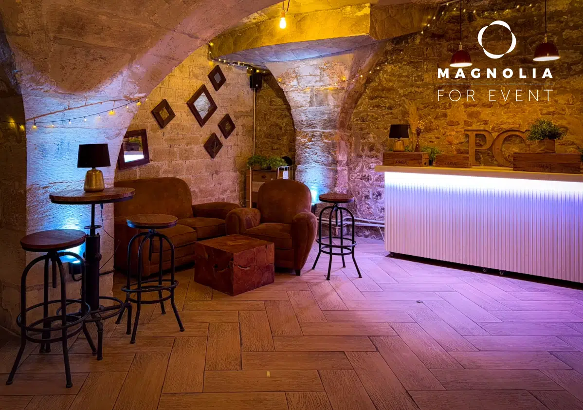 Salon confortable au PETIT PARIS CACHÉ (75004) avec des murs en pierre, un plafond voûté, un éclairage chaleureux, des fauteuils bruns, des petites tables, des tabourets de bar, un bar lumineux, des œuvres d'art encadrées et des guirlandes lumineuses. Logo Magnolia For Event dans le coin.