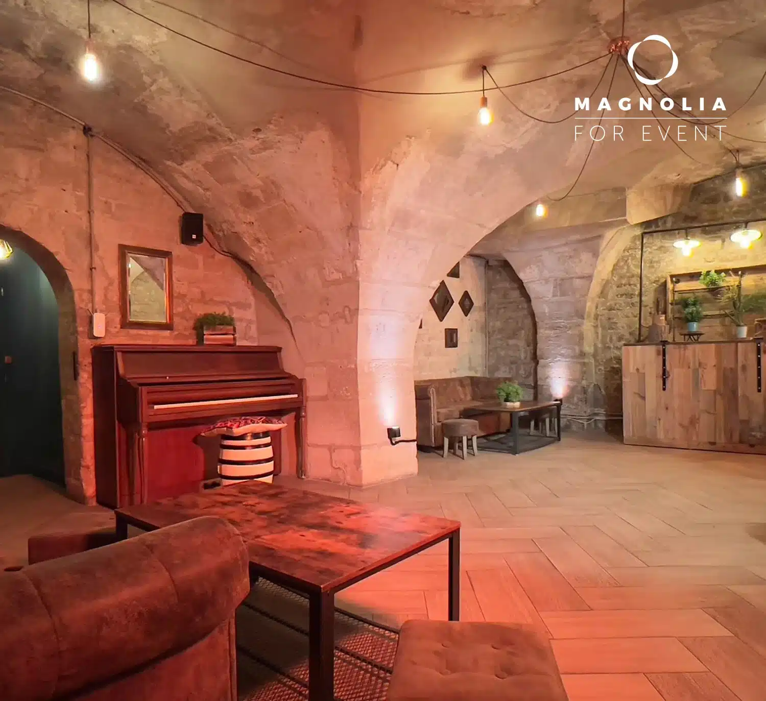LE PETIT PARIS CACHÉ (75004) propose un espace événementiel confortable avec des plafonds voûtés en pierre, un piano en bois, un canapé marron, une table, un éclairage chaleureux et des plantes en pot. Le logo Magnolia For Event est affiché dans le coin supérieur droit.